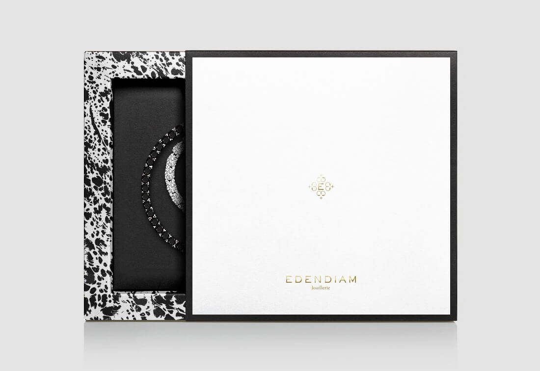 Une boîte à bijoux de luxe à bord moucheté blanc et noir, parfaite pour la création de catalogue, est partiellement ouverte pour révéler une pièce en forme de croissant noir et argenté à l'intérieur. La boîte porte l'inscription "EDENDIAM Joaillerie" en lettres d'or.