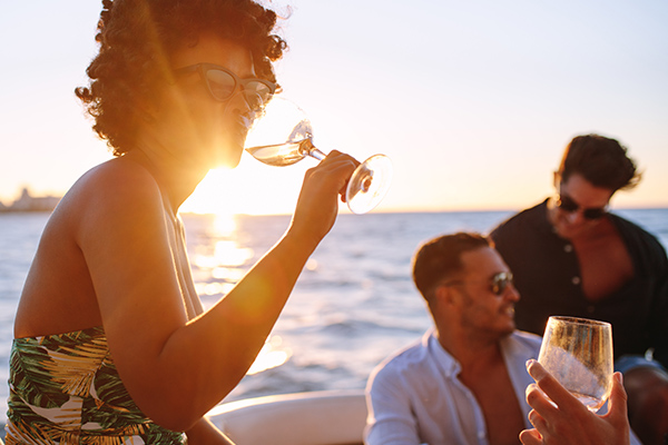 Trois personnes prennent un verre sur un bateau au coucher du soleil. L'une d'elles, portant des lunettes de soleil, sirote du vin tandis que les autres se détendent et discutent de leur dernier projet d'agence marketing digitale à Nice, avec l'océan et la lumière dorée du soleil en arrière-plan.