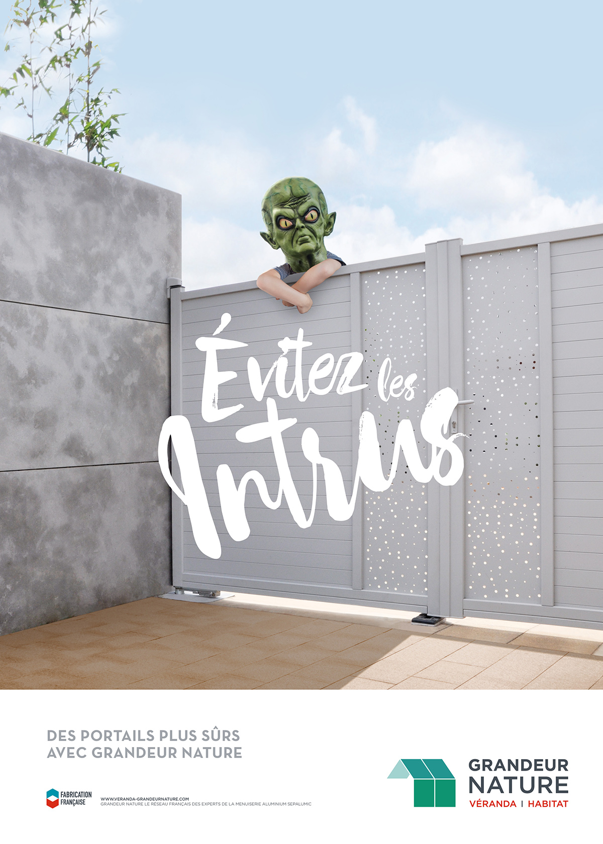 Une personne portant un masque de gobelin vert regarde par-dessus une clôture grise moderne sur laquelle est écrit en gros caractères blancs "Évitez les Intrus". Le texte ci-dessous fait la promotion des portails sécurisés et des solutions de réseau de communication de Grandeur Nature Véranda Habitat.