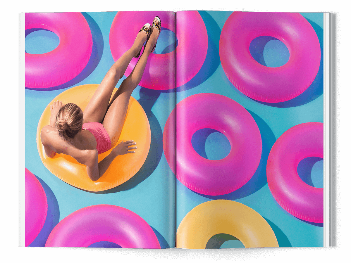 Une femme en maillot de bain rose se prélasse sur un flotteur de piscine jaune, entourée d'anneaux gonflables roses et jaunes sur un fond bleu vif, photographiée dans le cadre d'une vibrante création de catalogue dans un magazine ouvert.