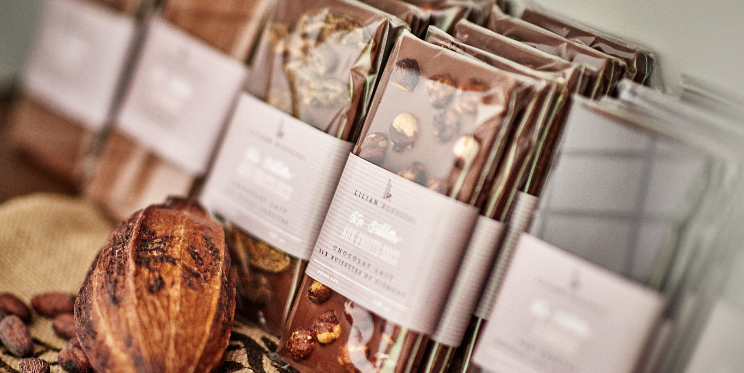 Plusieurs tablettes de chocolat dans un emballage transparent sont soigneusement disposées à côté d'une cabosse et de fèves de cacao entières sur une surface en toile de jute, ce qui en fait une installation idéale pour la création d'un catalogue avec des noix visibles et des étiquettes détaillées.