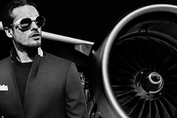 Un homme portant des lunettes de soleil et un manteau sombre se tient avec assurance à côté d'un moteur à réaction, incarnant une image de marque audacieuse. L'image en noir et blanc ajoute une touche élégante et dramatique, parfaite pour un marketing percutant.