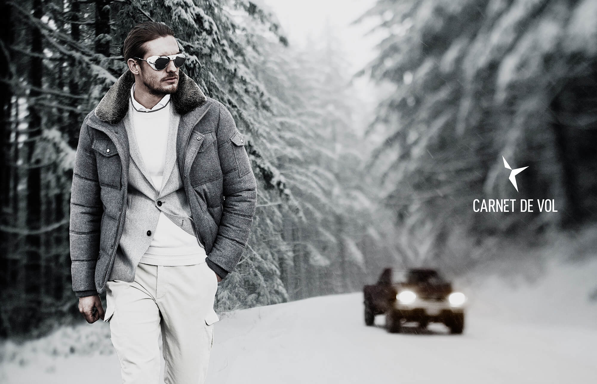 Un homme portant un manteau d'hiver gris, un col roulé blanc et des lunettes de soleil marche sur une route forestière enneigée. Un véhicule tout-terrain foncé roule à l'arrière-plan, mettant en évidence le marketing audacieux de CARNET DE VOL sur le côté droit.