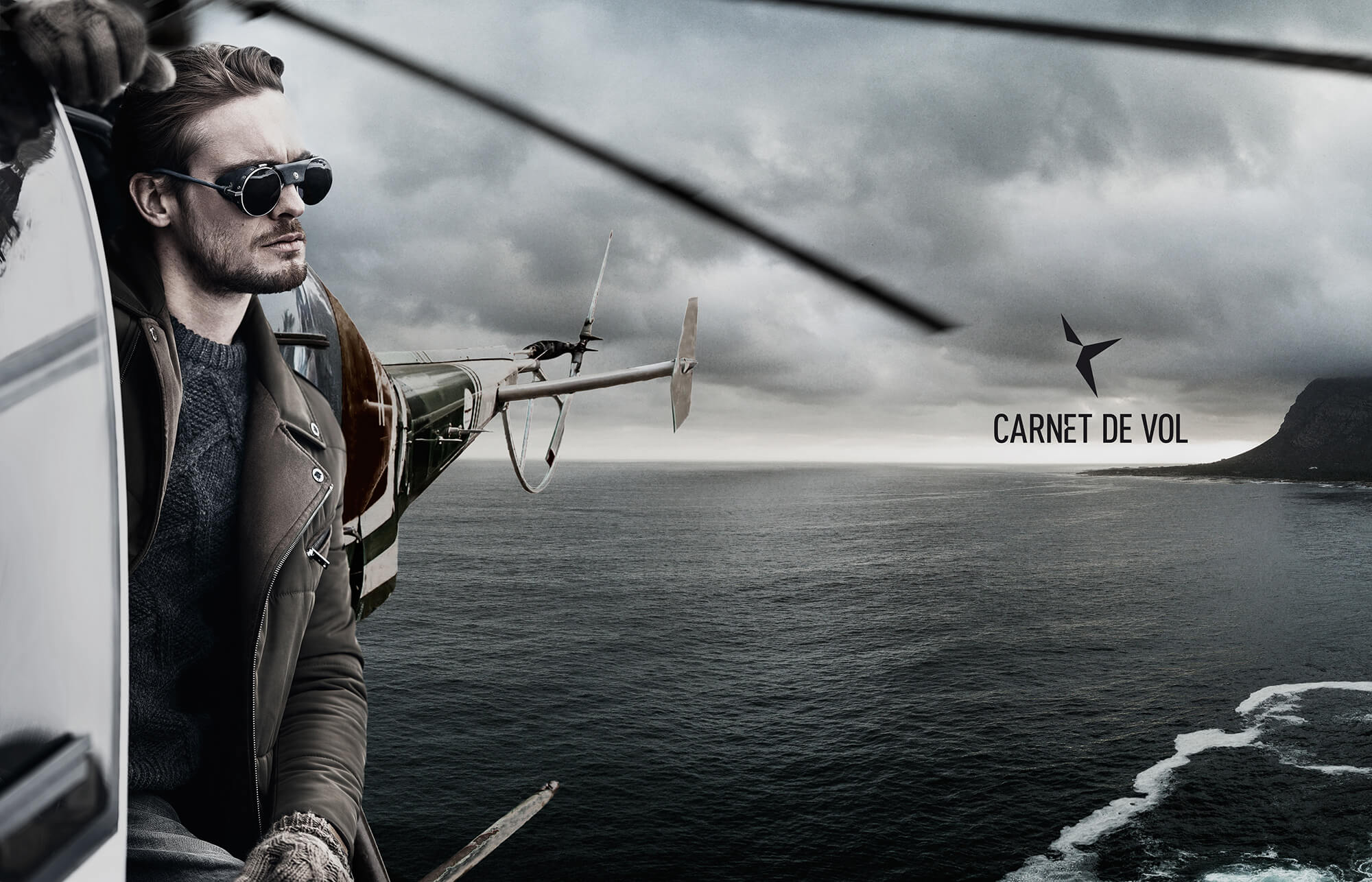 Un homme portant des lunettes de soleil et une veste se penche hors d'un hélicoptère volant au-dessus de l'océan, avec des nuages spectaculaires au-dessus de sa tête. Le logo "CARNET DE VOL" à droite met en évidence l'audace de la marque et du marketing en action.