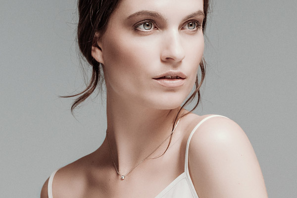 Une femme à la peau et aux yeux clairs regarde sur le côté, ses cheveux bruns étant coiffés librement. Portant un collier délicat et un caraco blanc, elle semble prête pour une création de catalogue sur un fond gris uni.