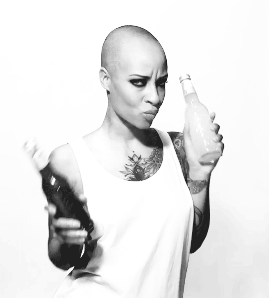 Une personne chauve, tatouée et aux yeux maquillés, tient deux bouteilles, montrant l'emballage tout en portant un débardeur blanc et en faisant une expression faciale enjouée sur un fond blanc uni.