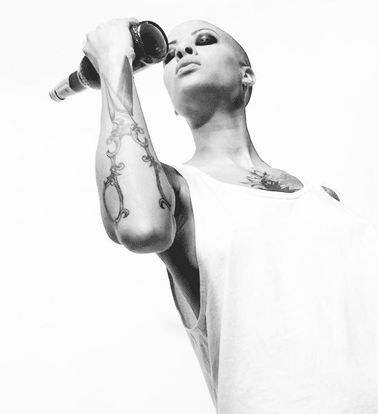 Une personne au crâne rasé, aux yeux maquillés, tatouée sur le bras et la poitrine, vêtue d'un haut blanc sans manches, tient un micro près de sa bouche tout en regardant vers le haut sur un fond blanc uni - comme un emballage audacieux pour une émotion brute.