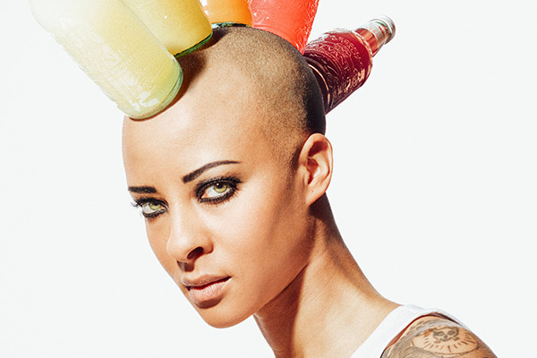 Une personne au crâne rasé et aux yeux très maquillés porte sur la tête des bouteilles d'emballage colorées disposées en mohawk. Elle porte un haut blanc et a un tatouage sur l'épaule. L'arrière-plan est blanc.