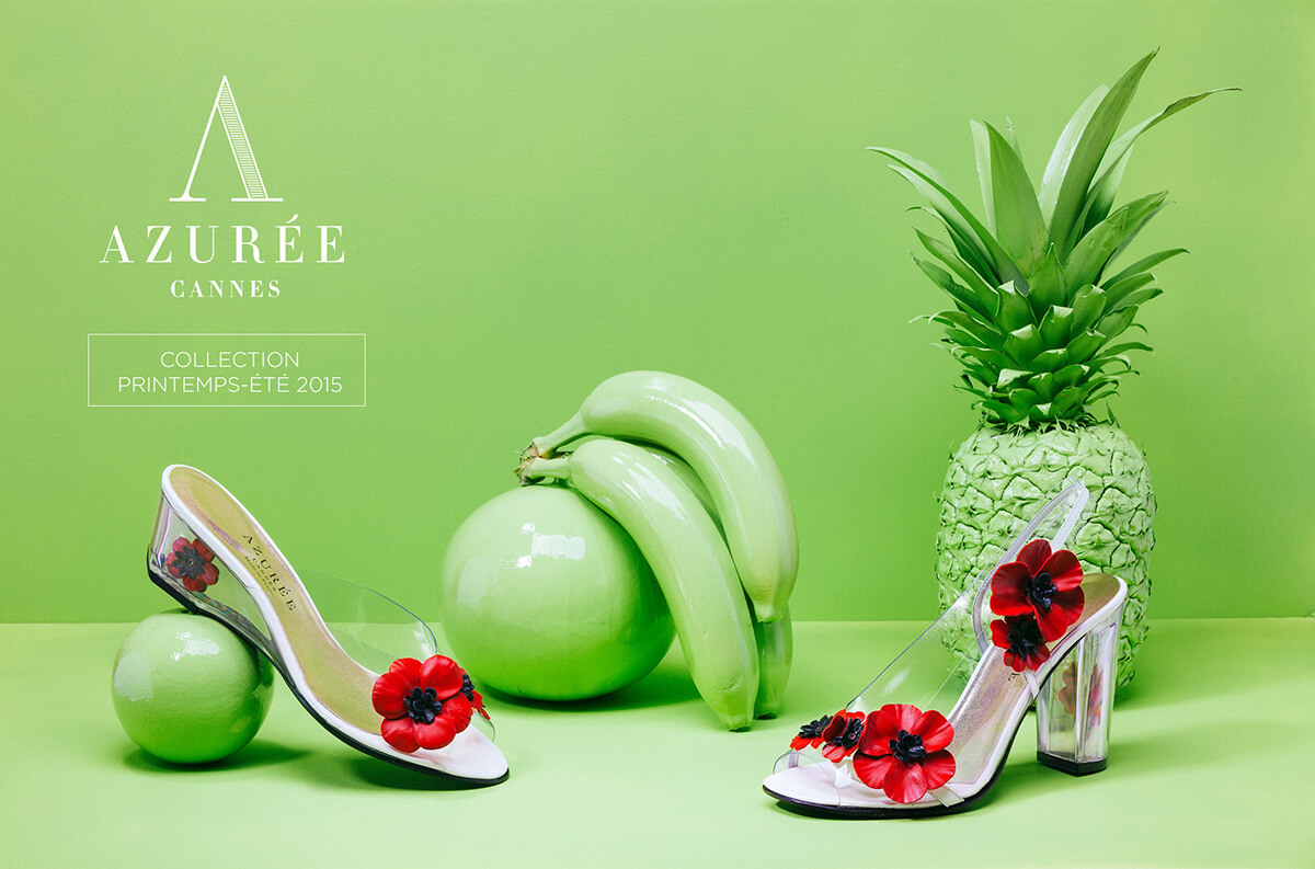 Deux chaussures à talon transparent avec des décorations de fleurs rouges sont présentées devant un fond vert, aux côtés d'accessoires de fruits vibrants, dans cette création de catalogue pour la collection AZURÉE CANNES Printemps-Été 2015.