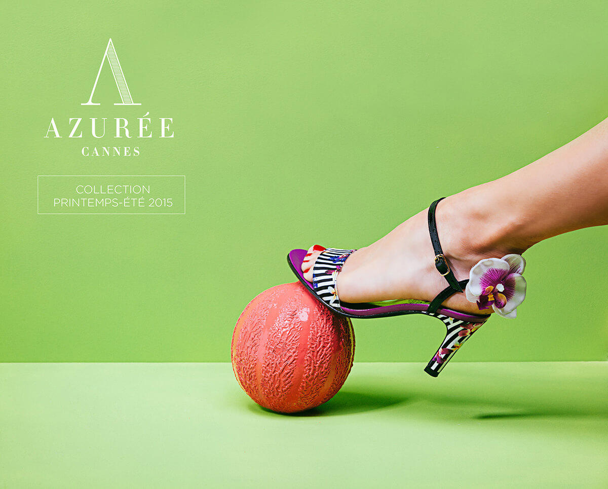 Le pied d'une femme portant une sandale à talon haut colorée avec un détail de fleur repose sur une balle texturée orange sur un fond vert, capturant l'essence de la création de catalogue pour l'Azurée Cannes Collection Printemps-Été 2015.