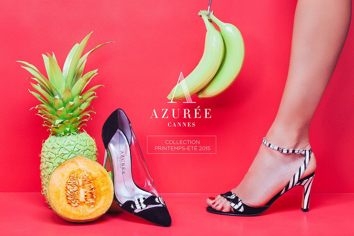 Une personne portant une chaussure à talon haut noire et blanche se tient à côté d'une chaussure assortie, de fruits et d'un texte vibrant sur fond de rose vif - évoquant l'art de la création de catalogue pour l'Azurée Cannes Collection Printemps-Été 2015.