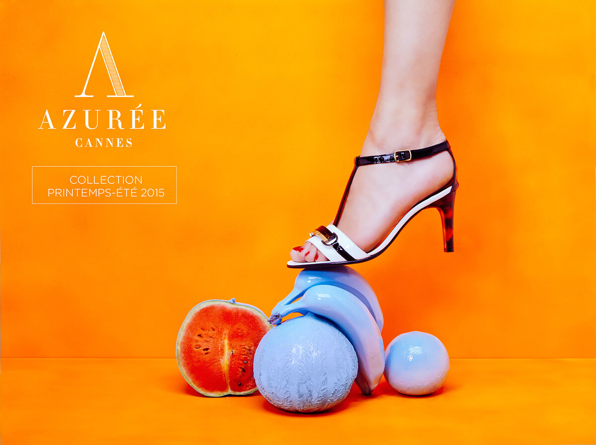 Le pied d'une femme chaussée d'une sandale à talon noire et blanche marche sur des fruits sculptés aux couleurs pastel sur un fond orange vibrant, mettant en valeur une création de catalogue pour AZURÉE CANNES, Collection Printemps-Été 2015.