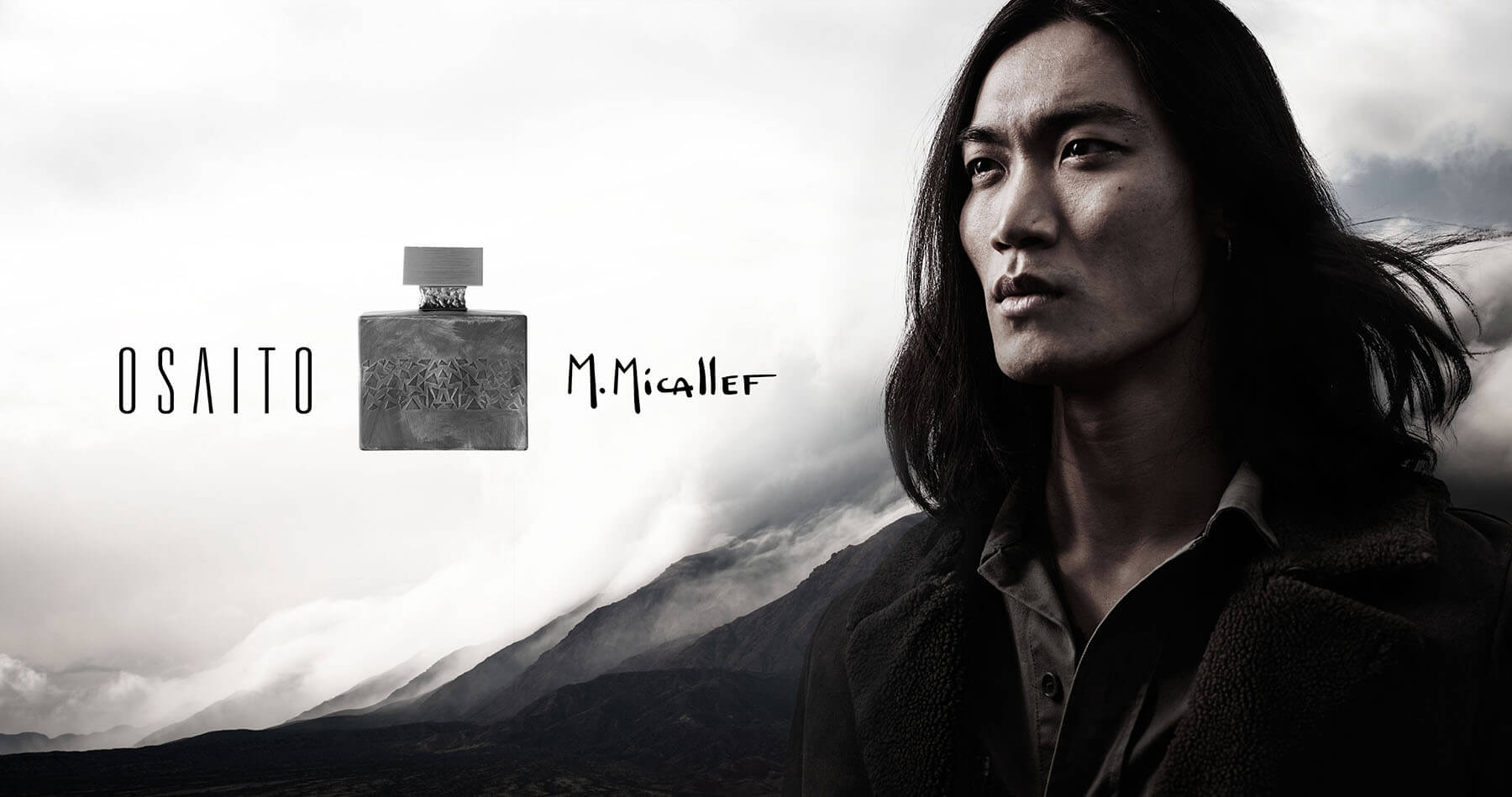 Un homme aux longs cheveux noirs se tient à l'extérieur sur fond de nuages et de montagnes spectaculaires, à côté d'un flacon de parfum OSAITO M. Micallef - une scène sombre et mystérieuse parfaite pour toute création de catalogue.