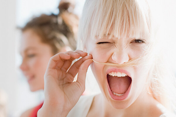 Une jeune femme enjouée aux cheveux blonds se fait une fausse moustache avec ses cheveux, en faisant un clin d'œil et en ouvrant grand la bouche - parfait pour une création de catalogue. Une autre femme brune est floue à l'arrière-plan. L'image est éclairée par une lumière claire et naturelle.