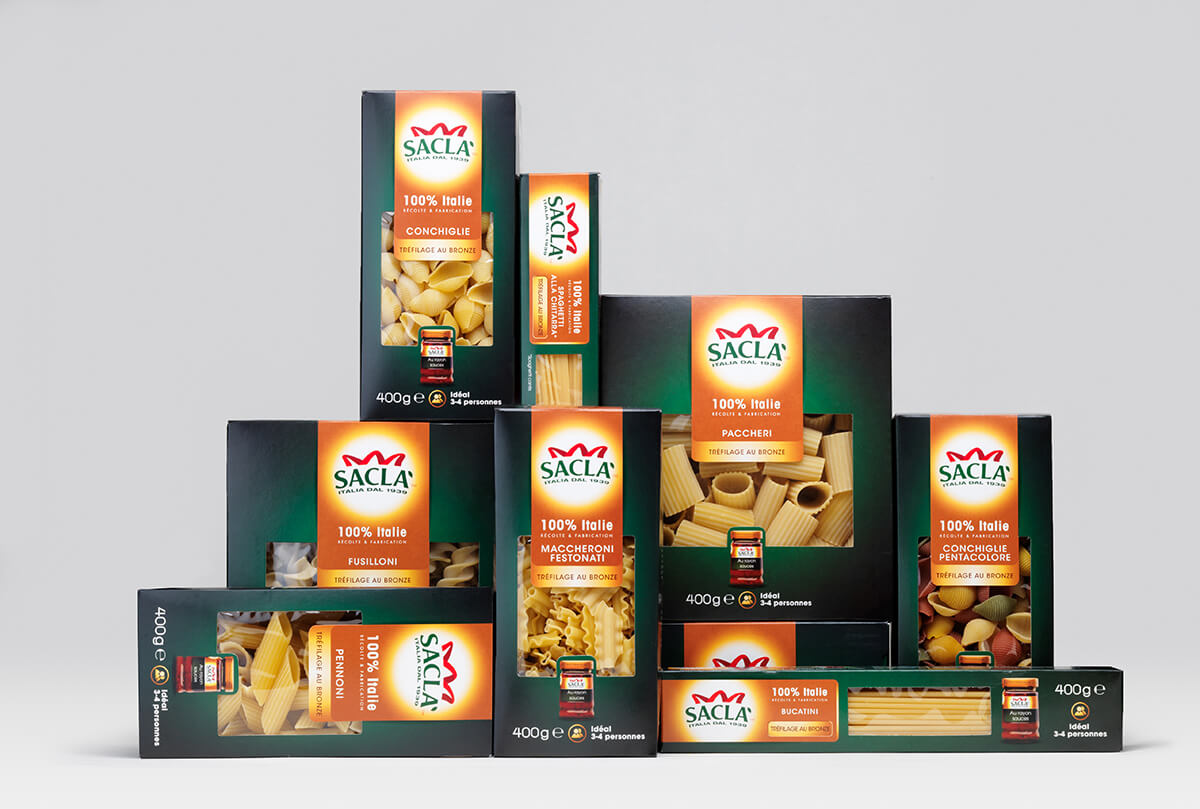Huit boîtes de pâtes italiennes de la marque Saclà, dans un emballage élégant, sont disposées en pyramide sur un fond gris uni. Chaque boîte noire comporte une fenêtre présentant les formes et les tailles de pâtes qu'elle contient.