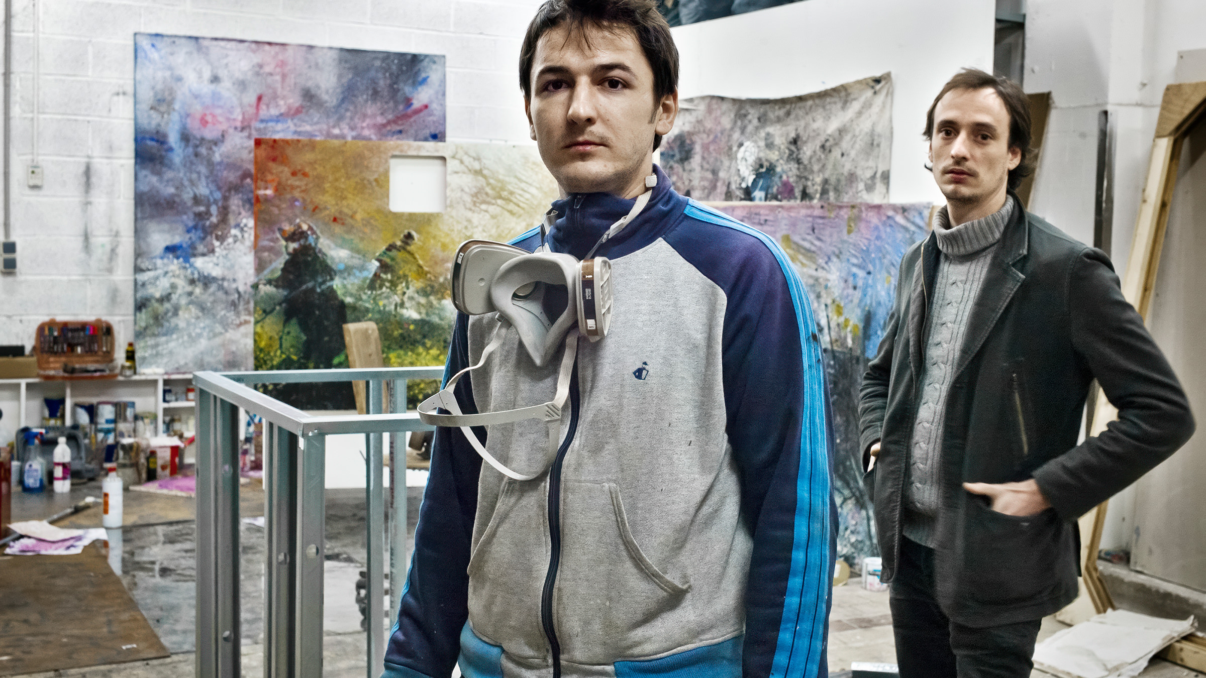 Deux hommes se tiennent dans un atelier d'art rempli de peintures abstraites colorées, discutant peut-être d'une création de livre. L'homme à l'avant porte un sweat à capuche gris et un masque respiratoire autour du cou ; l'homme à l'arrière porte un pull et un manteau. Des fournitures artistiques encombrent l'espace animé.