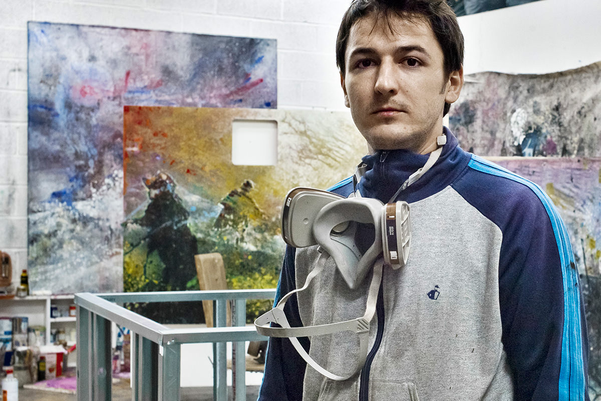 Un homme est debout à l'intérieur, vêtu d'un pull et d'un masque pour peindre, peut-être en envisageant une création de livre. Des objets abstraits ornent le mur derrière lui dans cette image colorée.