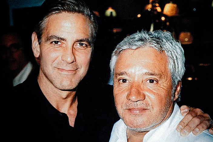 Deux hommes posent l'un près de l'autre pour une photo, tous deux légèrement souriants. L'un a les cheveux noirs et porte une chemise noire, tandis que l'autre a les cheveux gris courts et porte une chemise blanche - peut-être des collègues d'une agence web à Cannes. L'arrière-plan est faiblement éclairé.