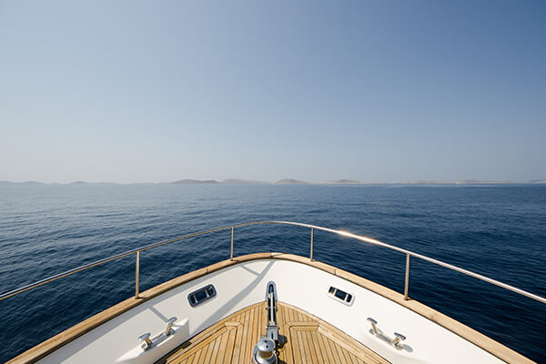 La proue d'un yacht, ornée d'un logo et d'une charte graphique épurés, navigue sur une eau calme et bleue, sous un ciel clair, avec des îles lointaines visibles à l'horizon.