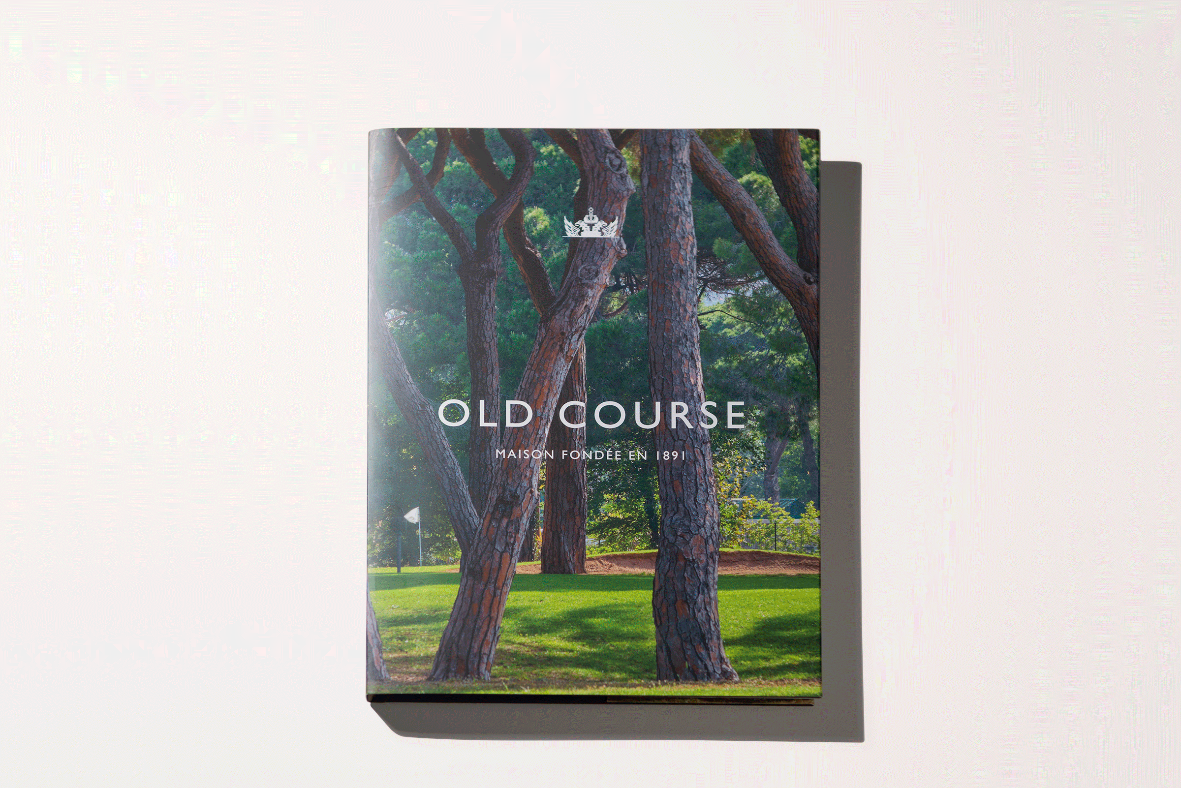 Un livre intitulé "OLD COURSE MAISON FONDÉE EN 1891" présente en couverture une photo de grands pins sur un terrain de golf, avec de l'herbe verte et un drapeau blanc - une image de marque reflétant l'élégance classique et la tradition sportive.
