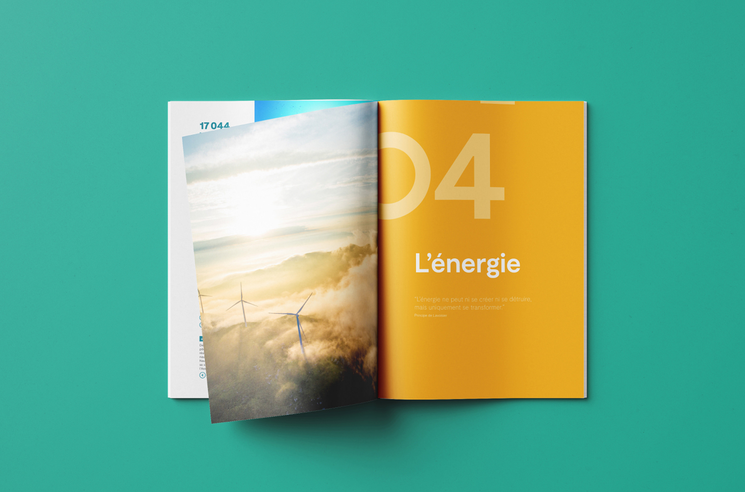 Un magazine rapport activité ouvert présente des éoliennes dans un paysage brumeux sur une page, et un fond jaune avec "04" en gras et le mot français "L'énergie" sur l'autre page. Le magazine repose sur une surface sarcelle.