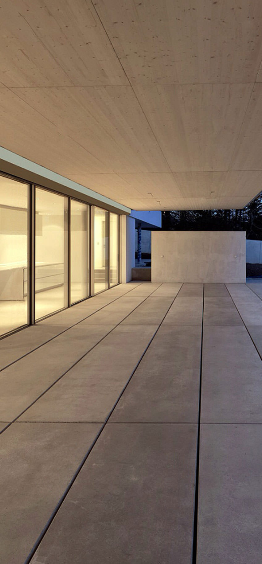 Un patio moderne et minimaliste avec de grandes dalles de béton et un plafond en bois, conçu par une agence de communication BTP, comporte des portes vitrées allant du sol au plafond et donnant accès à un espace intérieur très éclairé à la tombée de la nuit.