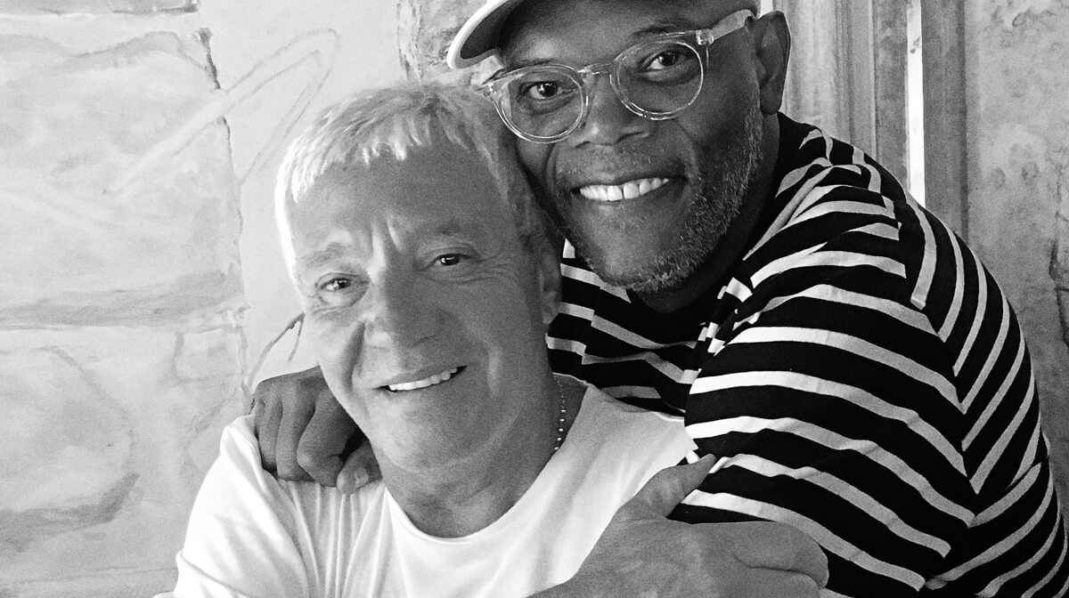 Deux hommes sourient et posent ensemble pour une photo. L'un, portant une chemise rayée, des lunettes et une casquette, serre dans ses bras l'autre homme aux cheveux courts et clairs et à la chemise unie. Capturés en noir et blanc, ils ressemblent à des collègues d'une agence web à Cannes.