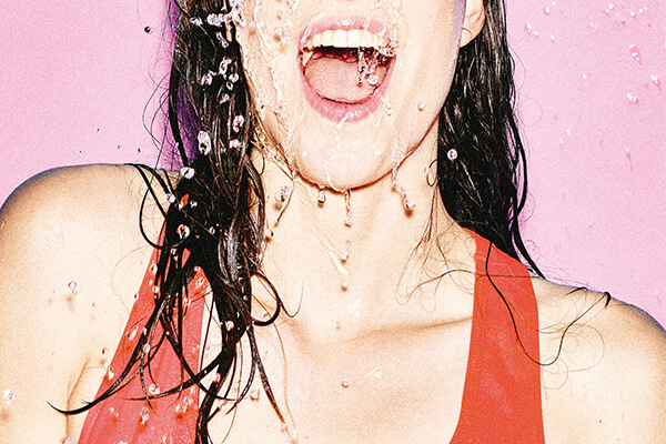 Une personne portant un haut rouge se tient sur un fond rose, les cheveux mouillés et la bouche ouverte, tandis que de l'eau éclabousse son visage et son cou, capturant ainsi la créativité audacieuse d'une campagne d'agence créative.