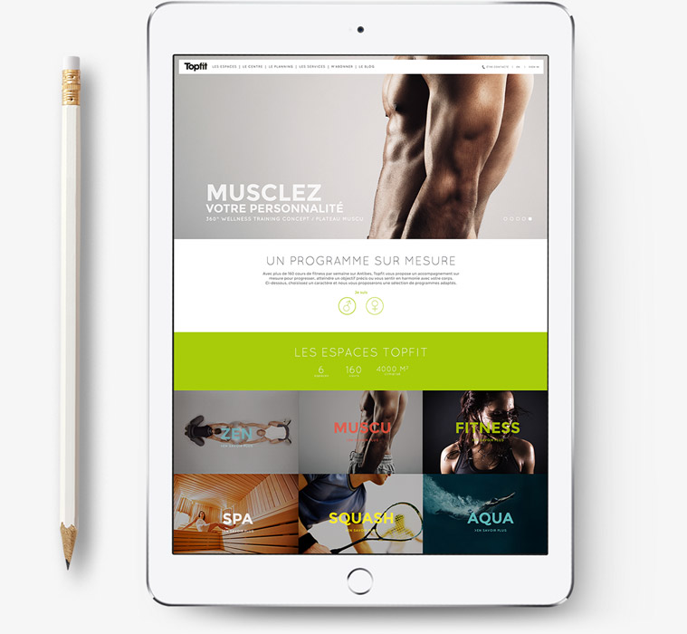 Une tablette affichant un site web de fitness avec une forte image de marque, présentant des images de bras musclés, différentes catégories d'entraînement et une section verte mettant en évidence divers programmes de fitness. Un crayon est posé à côté de la tablette sur une surface blanche.
