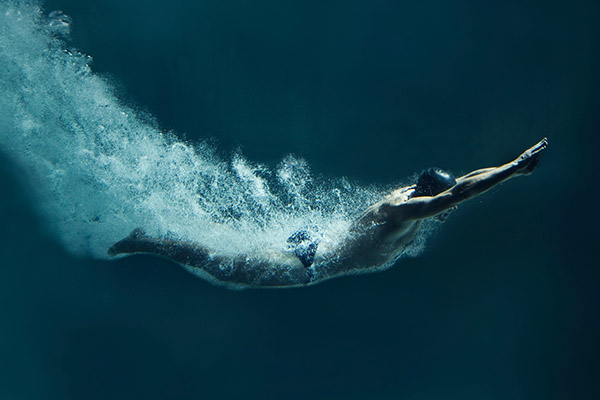 Un nageur vêtu d'un maillot de bain bleu, arborant une marque élégante, glisse sous l'eau les bras tendus vers l'avant, laissant une traînée de bulles derrière lui et créant une sensation dynamique de mouvement et de vitesse.