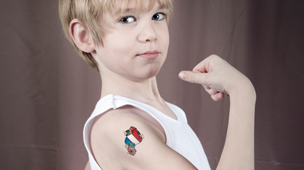 Un jeune garçon vêtu d'une chemise blanche sans manche fléchit le bras, montrant un tatouage temporaire de la carte de l'Italie avec les couleurs du drapeau italien - un clin d'œil ludique au marketing territorial et au branding - sur un fond marron uni.