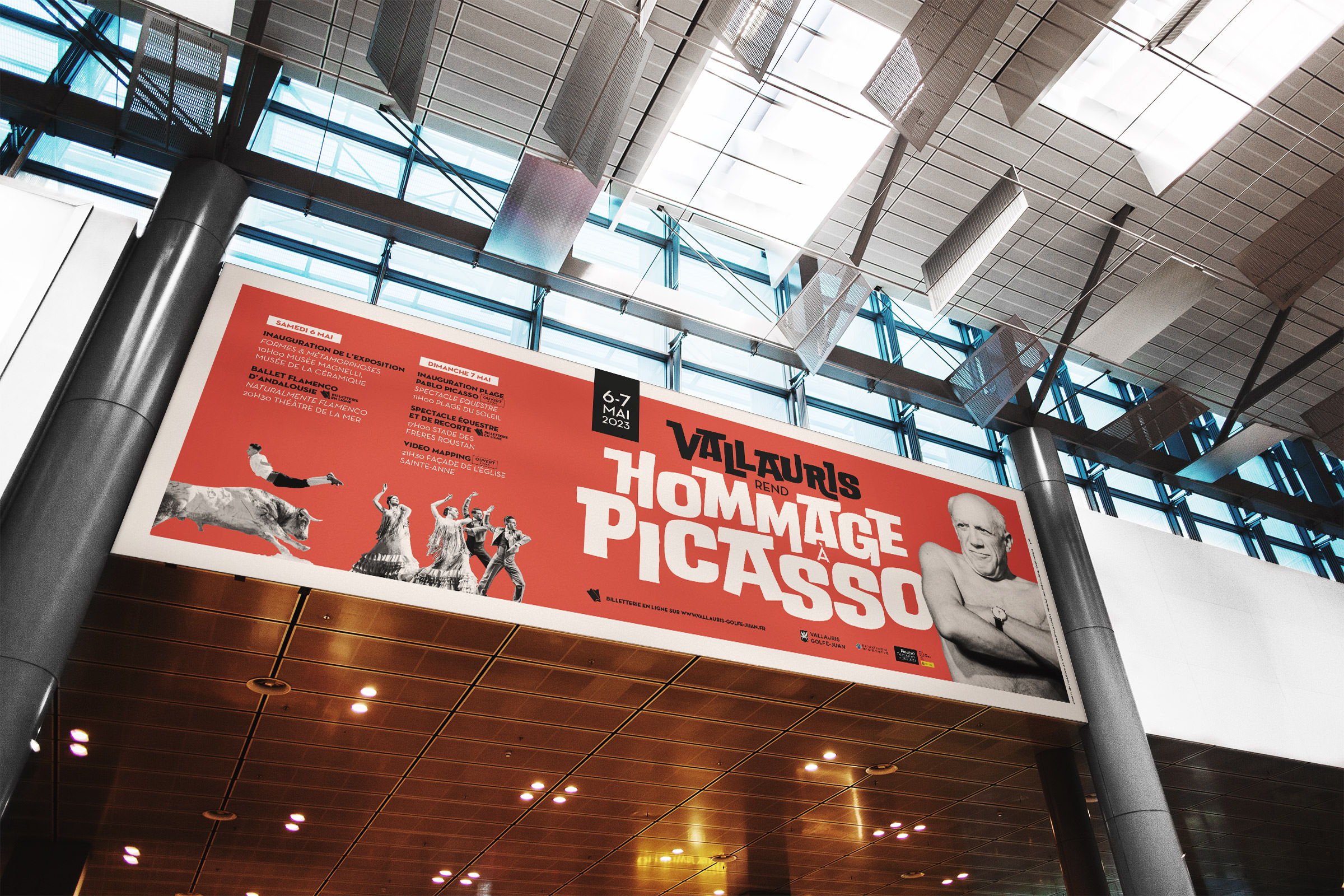 Grande banderole rouge à l'intérieur d'un bâtiment moderne avec des plafonds en verre, comportant le texte "Hommage à Picasso", les détails de l'événement, des photos de Picasso, des danseurs, un taureau et un pot - soulignant la communication événementielle pour un hommage à Picasso à Vallauris.