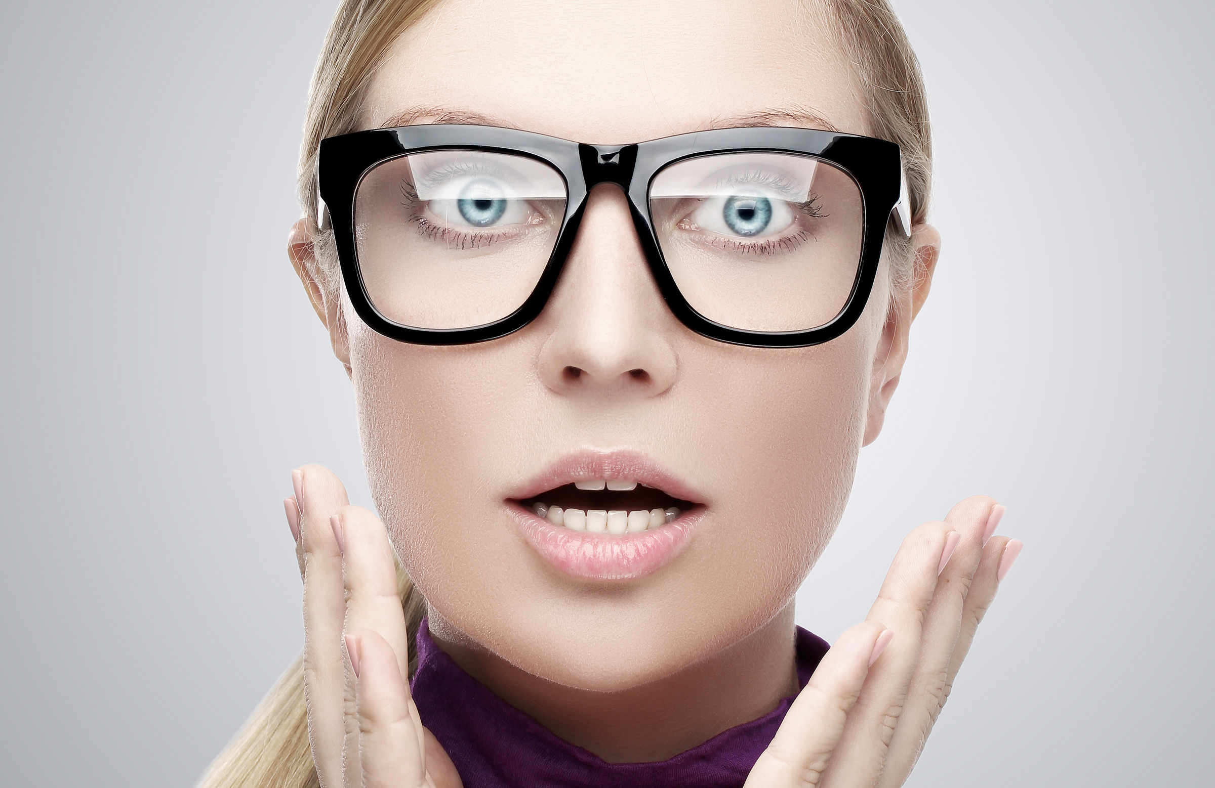 Une femme surprise, aux yeux écarquillés et à la bouche ouverte, porte de grosses lunettes noires. Ses mains sont levées près de son visage, elle a des cheveux blonds et un teint clair, sur un fond neutre.
