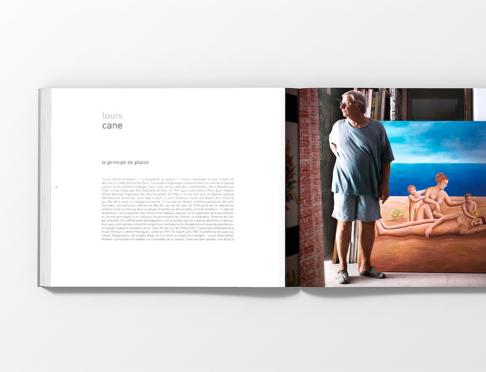 Un homme en tenue décontractée se tient à l'intérieur, à côté d'une grande peinture représentant des nus allongés sur une plage. Le livre ouvert, issu d'une édition spéciale, présente le texte sur la page de gauche et la photo sur la page de droite, sur un fond blanc.