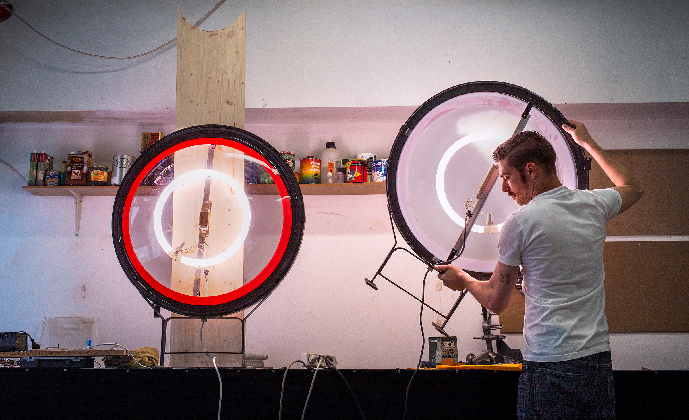 Un homme vêtu d'un tee-shirt blanc ajuste un luminaire circulaire sur un établi, avec un autre anneau lumineux à côté de lui - son installation concentrée reflète une précision similaire aux stratégies de webmarketing. Des étagères contenant des bocaux et des boîtes de conserve sont fixées au mur à l'arrière-plan.