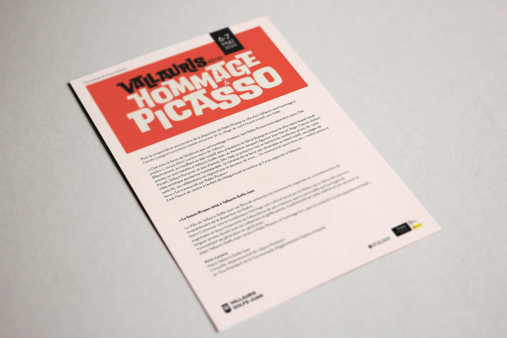 Un prospectus imprimé pour "Vallauris Hommage à Picasso" présente un texte rouge et noir en gras en haut et les détails de l'événement en bas sur un fond blanc, soulignant son utilisation dans les relations presse événementielles, couché de biais sur une surface de couleur claire.