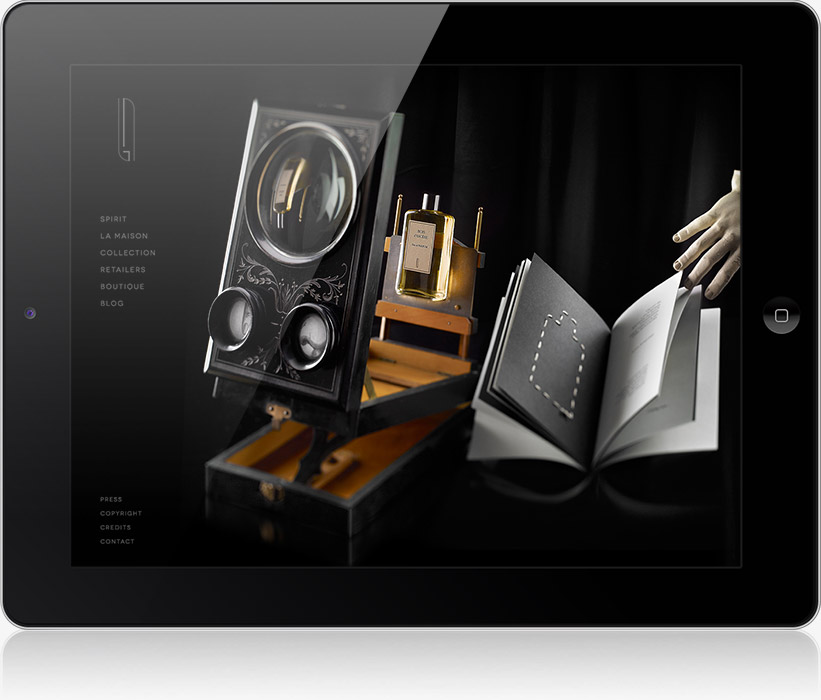 Une tablette présente un site web sombre et élégant d'une agence de communication digitale à Nice, avec un flacon de parfum de luxe dans un coffret décoratif, une main interagissant avec un livre ouvert et des liens de navigation élégants sur le côté gauche.