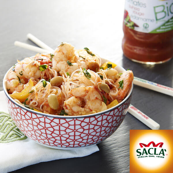 Un bol à motifs rempli de crevettes, de nouilles, de légumes et d'olives est posé sur une serviette avec des baguettes à proximité. Un pot de sauce tomate et le logo Saclà sont visibles à l'arrière-plan, soulignant l'importance accordée par Saclà à la saveur et à la gestion communautaire.