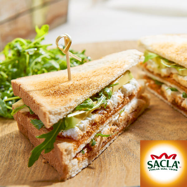 Deux sandwichs triangulaires grillés, garnis de légumes verts, de fromage et de légumes, servis sur une planche en bois et accompagnés de roquette fraîche - parfaits pour un déjeuner de Community Management. Le logo de la marque Saclà apparaît dans le coin inférieur droit.