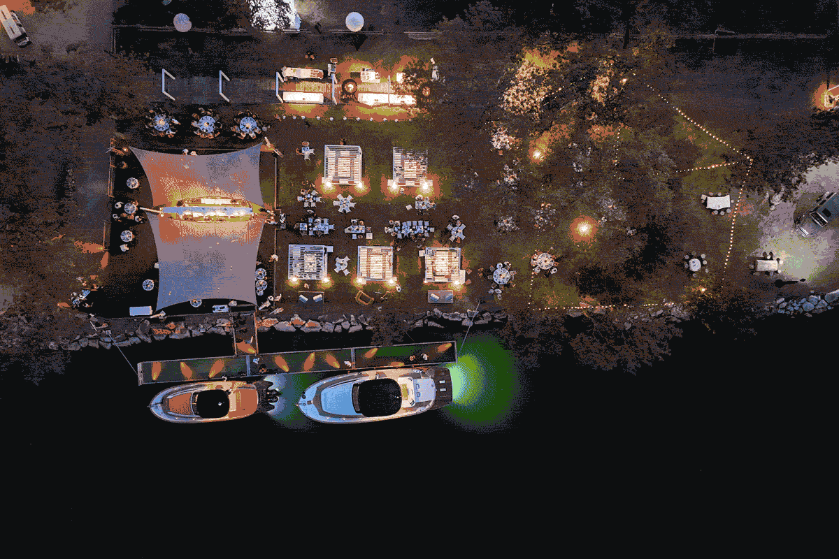 Vue aérienne d'un événement nocturne en plein air, au bord de l'eau, avec des tables décorées, des guirlandes lumineuses, des tentes et deux bateaux à quai éclairés par des lumières vertes et orange - un cadre idéal pour des moments uniques de branding produit, le tout entouré d'arbres.