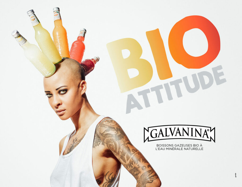 Une personne au crâne rasé pose avec assurance dans un débardeur blanc. Des bouteilles colorées sont disposées sur sa tête comme une couronne, mettant en valeur une publicité créative. Le texte "BIO ATTITUDE" en gros caractères et le logo Galvanina avec le slogan français apparaissent en dessous.