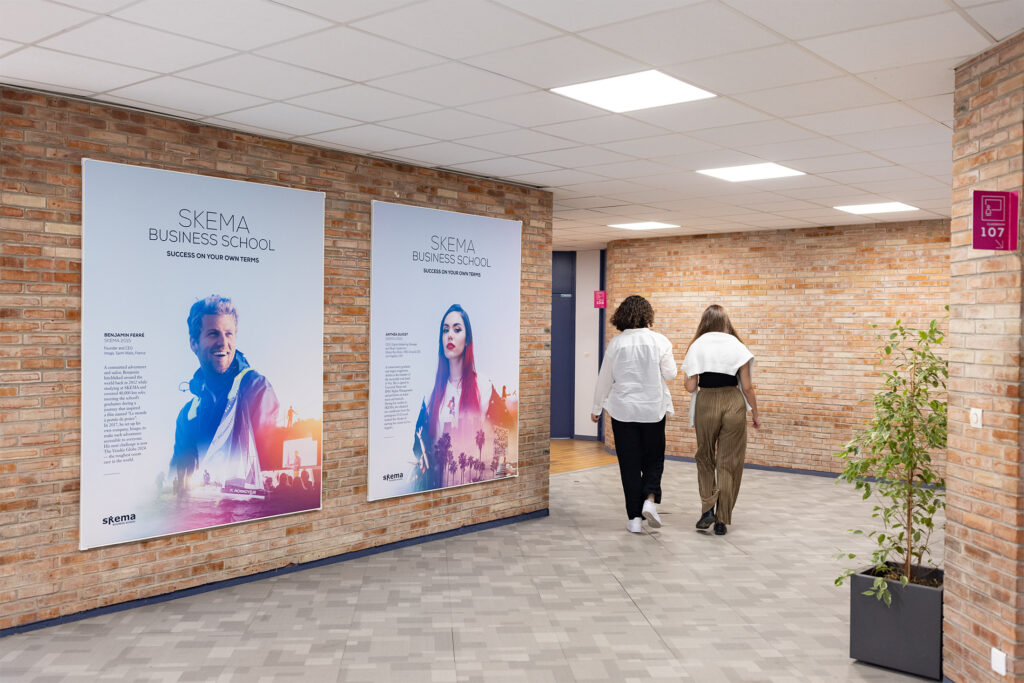 Deux personnes marchent dans un couloir aux murs de briques et passent devant de grandes affiches de la SKEMA Business School. Elles présentent le nom de l'école, son slogan et une pub de création frappante, ainsi qu'un homme et une femme à côté d'une plante en pot.