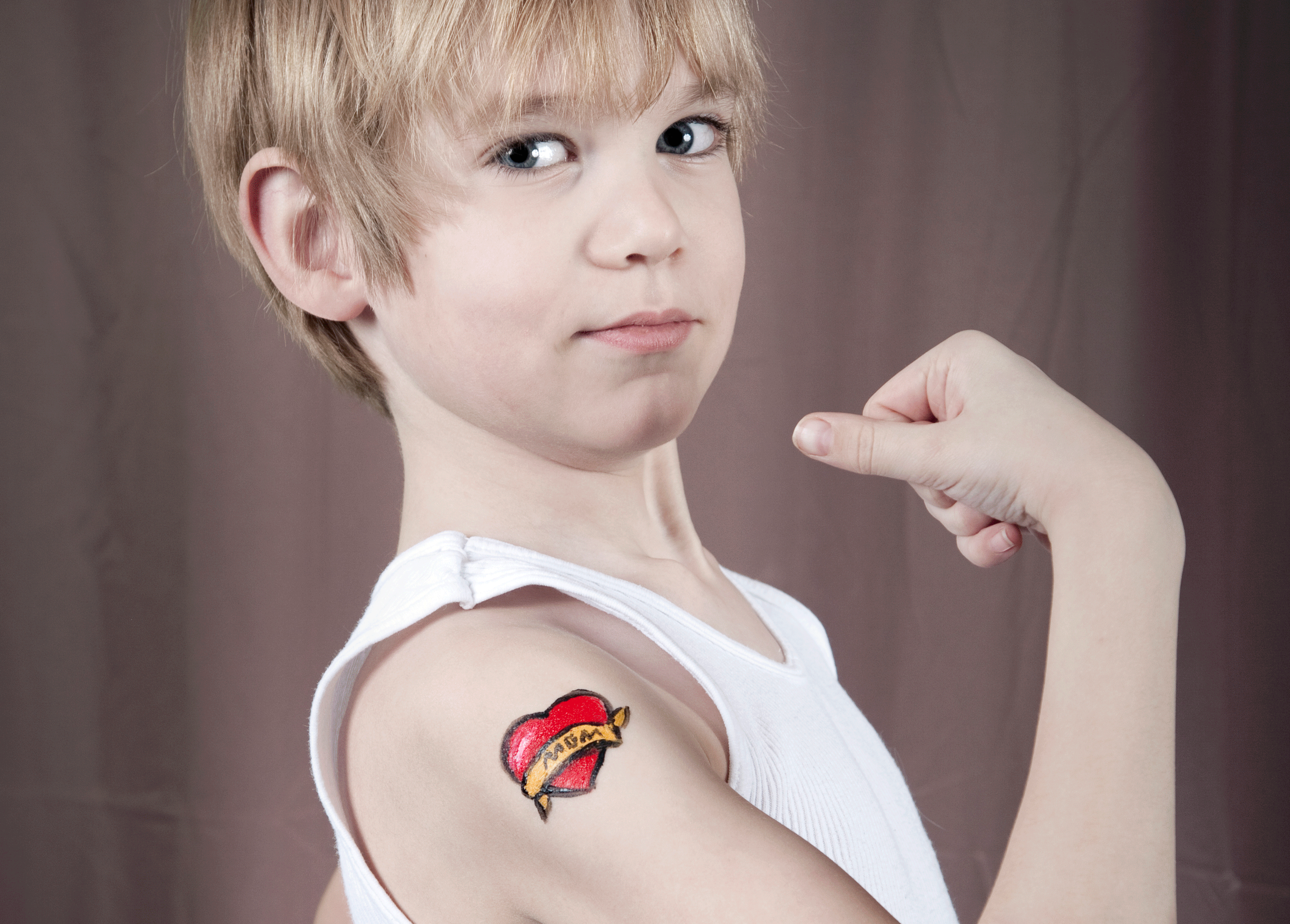 Un jeune garçon vêtu d'un débardeur blanc fléchit le bras et montre un tatouage temporaire représentant un cœur rouge avec une bannière jaune portant l'inscription "MOM" sur la partie supérieure de son bras, mettant en valeur son approche ludique de la marque personnelle sur un fond de rideau marron neutre.