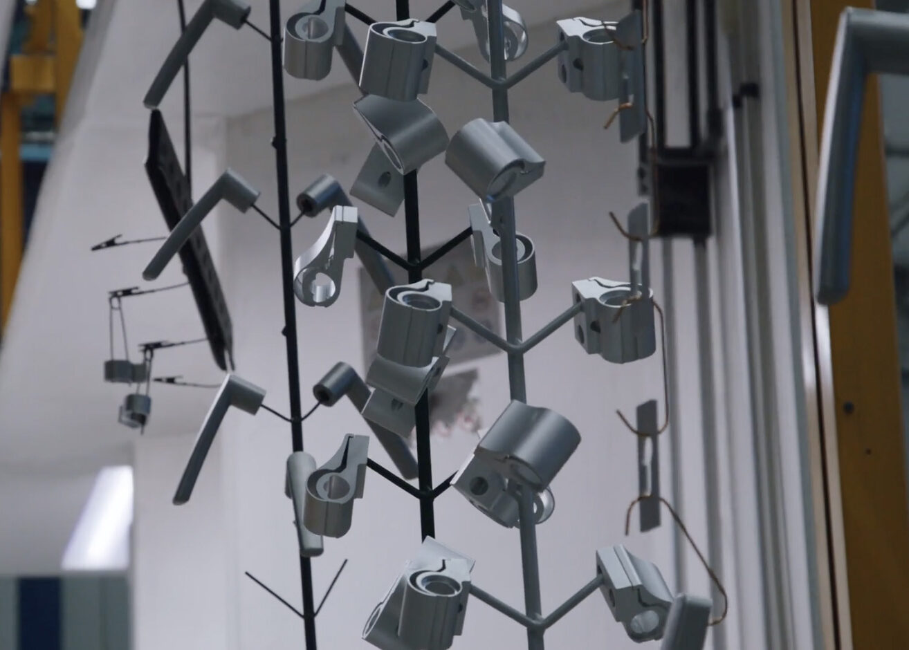 Des pièces métalliques sont montées sur des racks et suspendues verticalement, éventuellement pour être peintes ou revêtues, à l'intérieur d'une installation industrielle. Les équipes vidéo de l'Agence ont pu capturer les composants récemment fabriqués et régulièrement espacés dans des images de haute qualité.