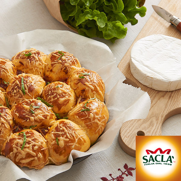 Un plat de petits pains dorés au fromage est posé sur du papier sulfurisé à côté d'une laitue verte fraîche et d'une meule de brie sur une planche en bois - parfait pour votre prochain événement de gestion communautaire. Le logo Saclà apparaît dans le coin inférieur droit.