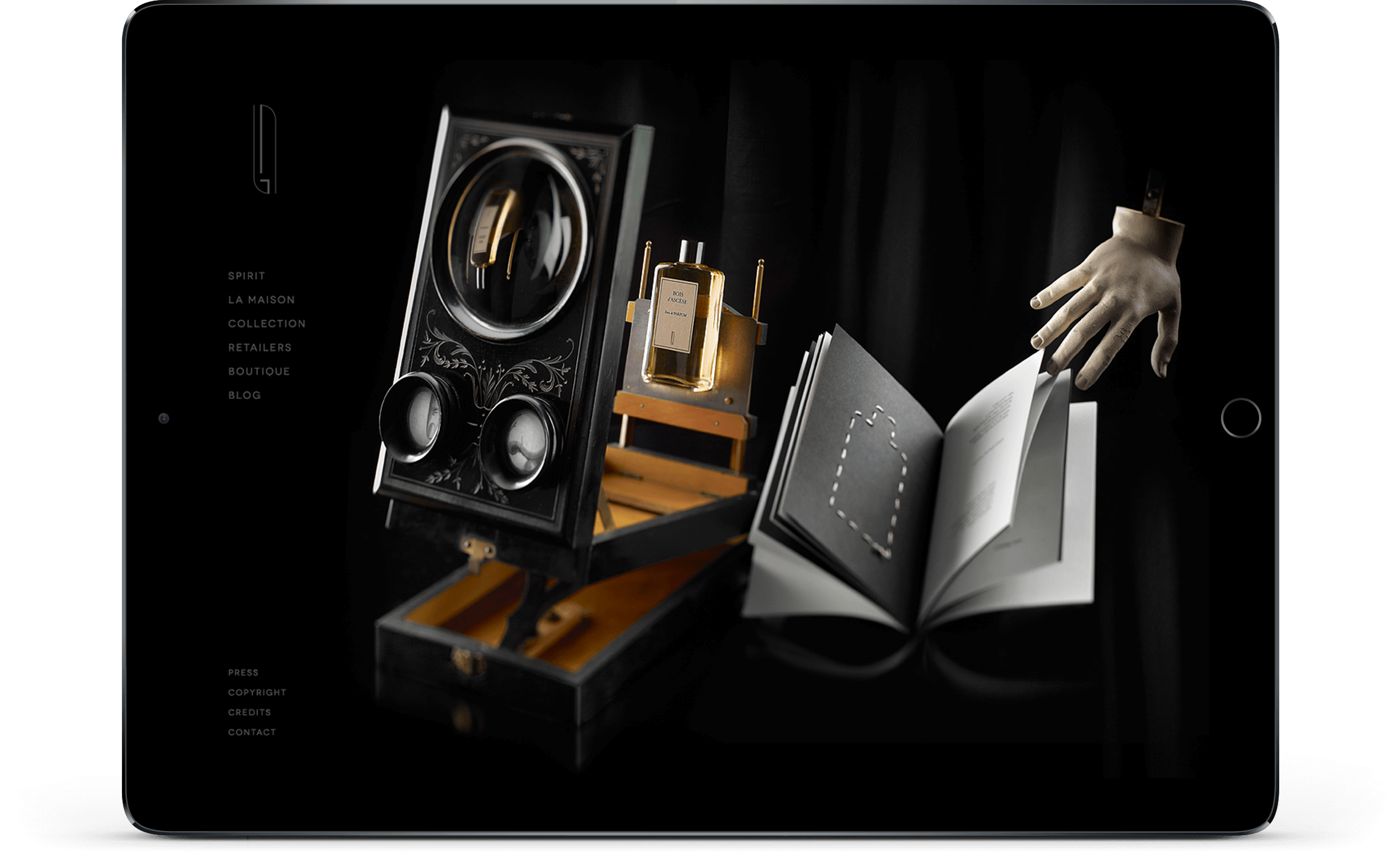 Un écran d'iPad affiche un site de création internet au thème sombre, avec une boîte de style vintage avec des cadrans, un livre ouvert, un flacon de parfum et une main de mannequin qui tend le livre. Des menus textuels minimaux apparaissent sur le côté gauche.