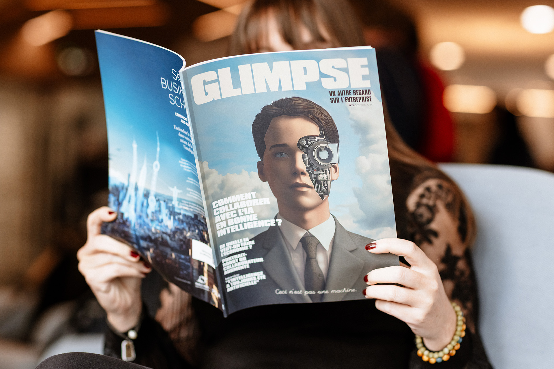 Une personne aux cheveux longs et aux ongles peints lit un magazine intitulé "GLIMPSE", dont la couverture présente un homme à l'œil robotisé. Le visage de la personne est caché derrière le magazine en papier glacé.