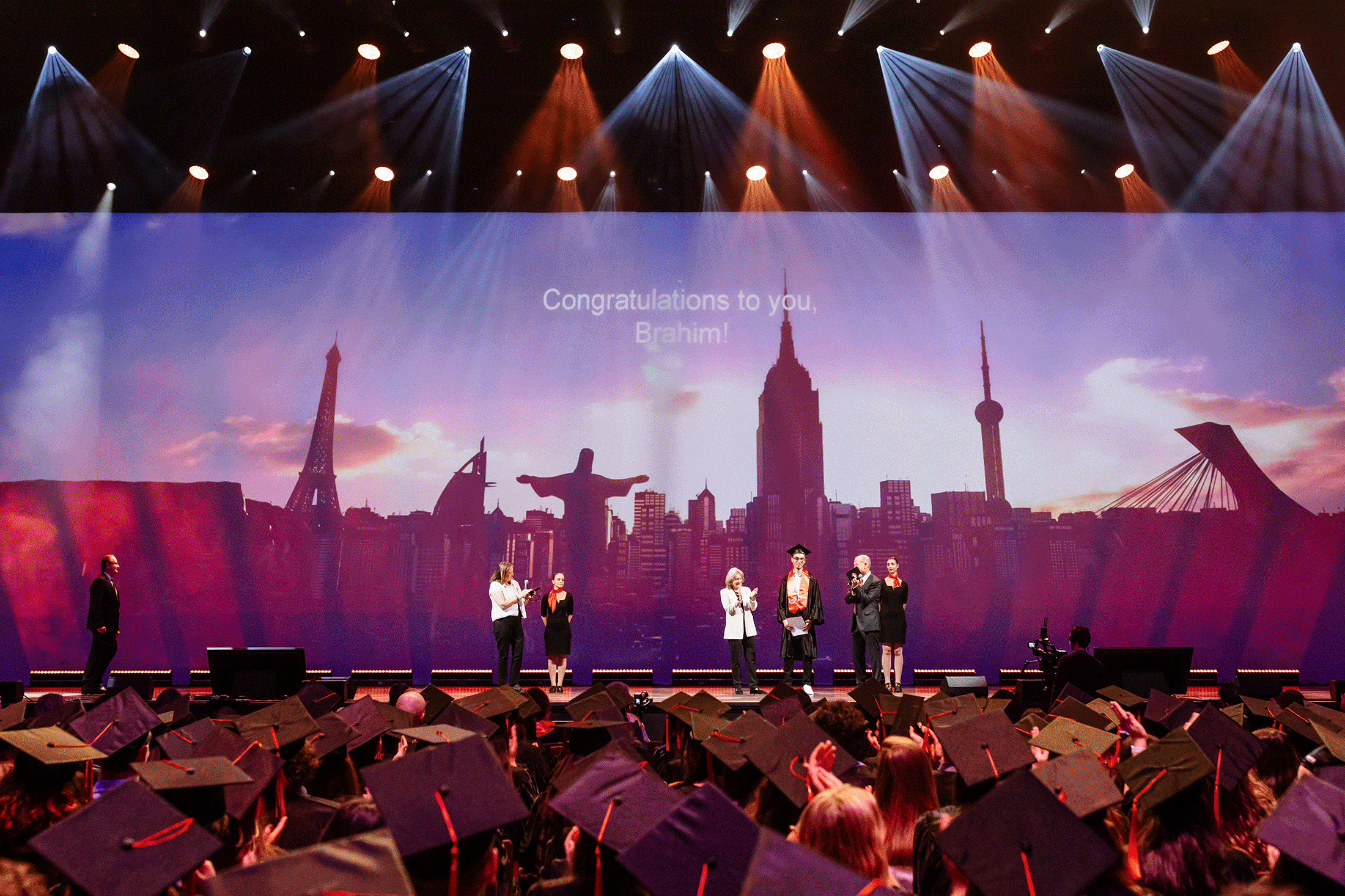 Cérémonie de remise de diplômes avec des diplômés portant des casquettes et faisant face à une scène où se tiennent plusieurs personnes sous des lumières vives. La communication événementielle d'un expert est mise en valeur. La toile de fond représente l'horizon d'une ville et l'on peut y lire : "Félicitations à toi, Brahim !.