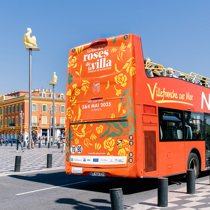 Un bus touristique à impériale rouge vif, orné d'une publicité événementielle florale et d'un message Création d'identité de marque, est stationné sur une place ensoleillée, près de grandes statues et de bâtiments colorés, sous un ciel bleu dégagé.