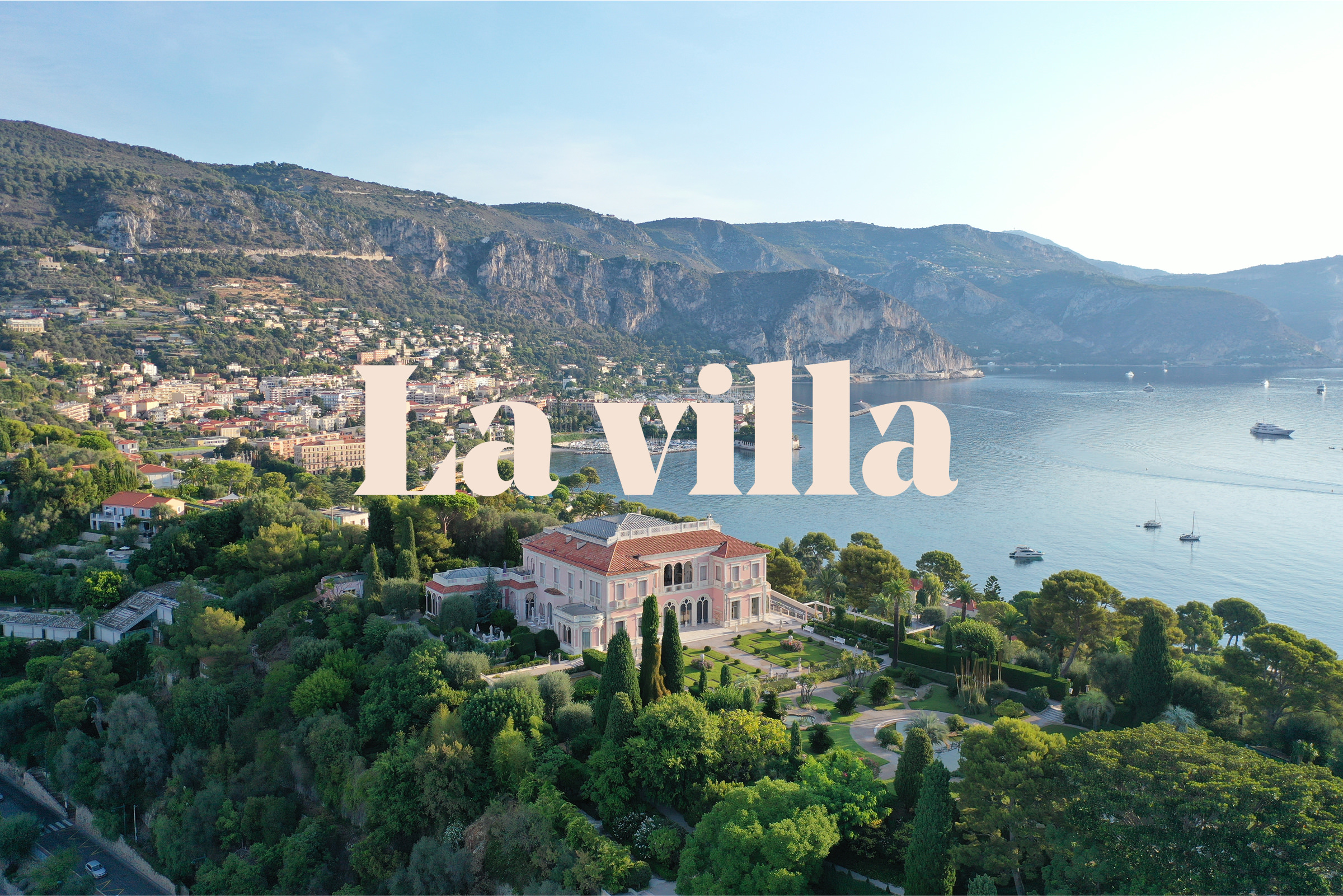 Vue aérienne d'une grande villa entourée de jardins luxuriants sur un littoral, avec des montagnes et une ville en arrière-plan. Les mots "La villa" sont superposés en grand texte pâle, reflétant la création d'identité de marque.